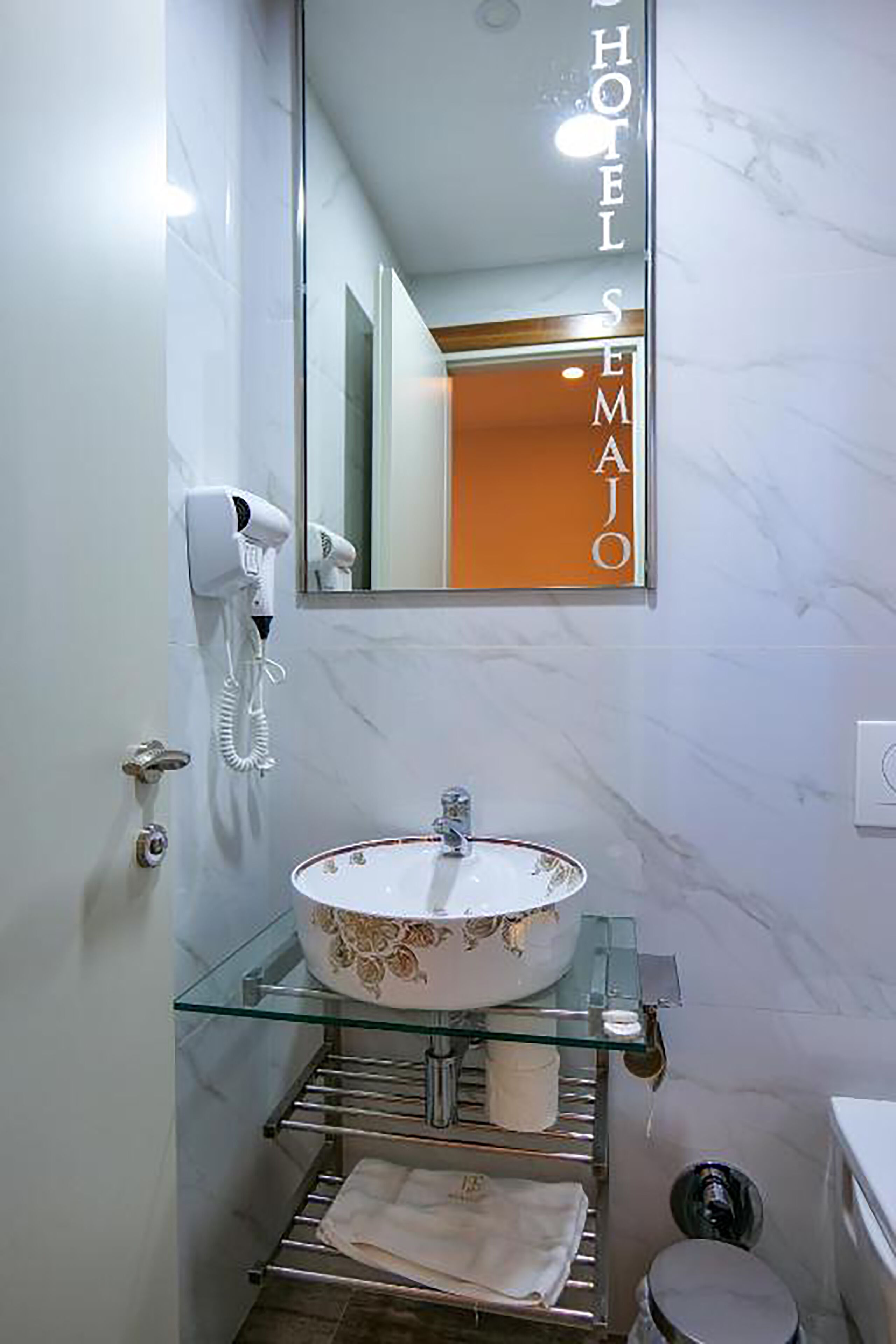A bathroom with a glass shelf holding toiletries, a white sink with a floral design, and a hairdryer mounted on the wall.