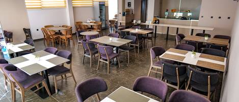 A modern dining area with multiple tables and chairs, a kitchen in the background, and a large mirror on the wall.