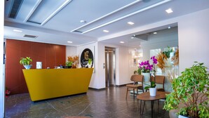 Zona de recepción de hotel con un mostrador amarillo, una mesa y un área de descanso con sillas.