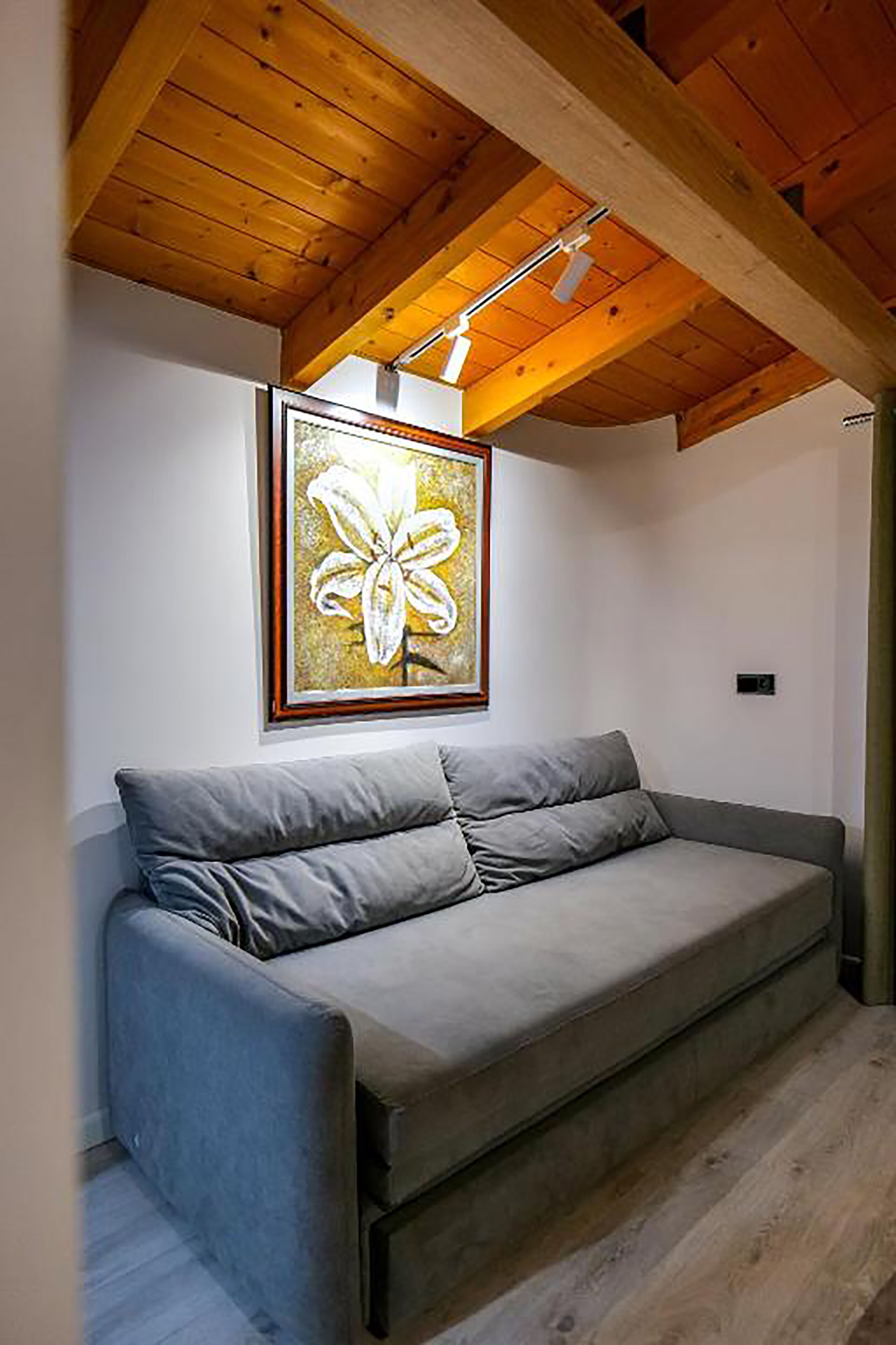 A modern living room with a grey sofa, wooden ceiling beams, and a framed artwork of a flower.