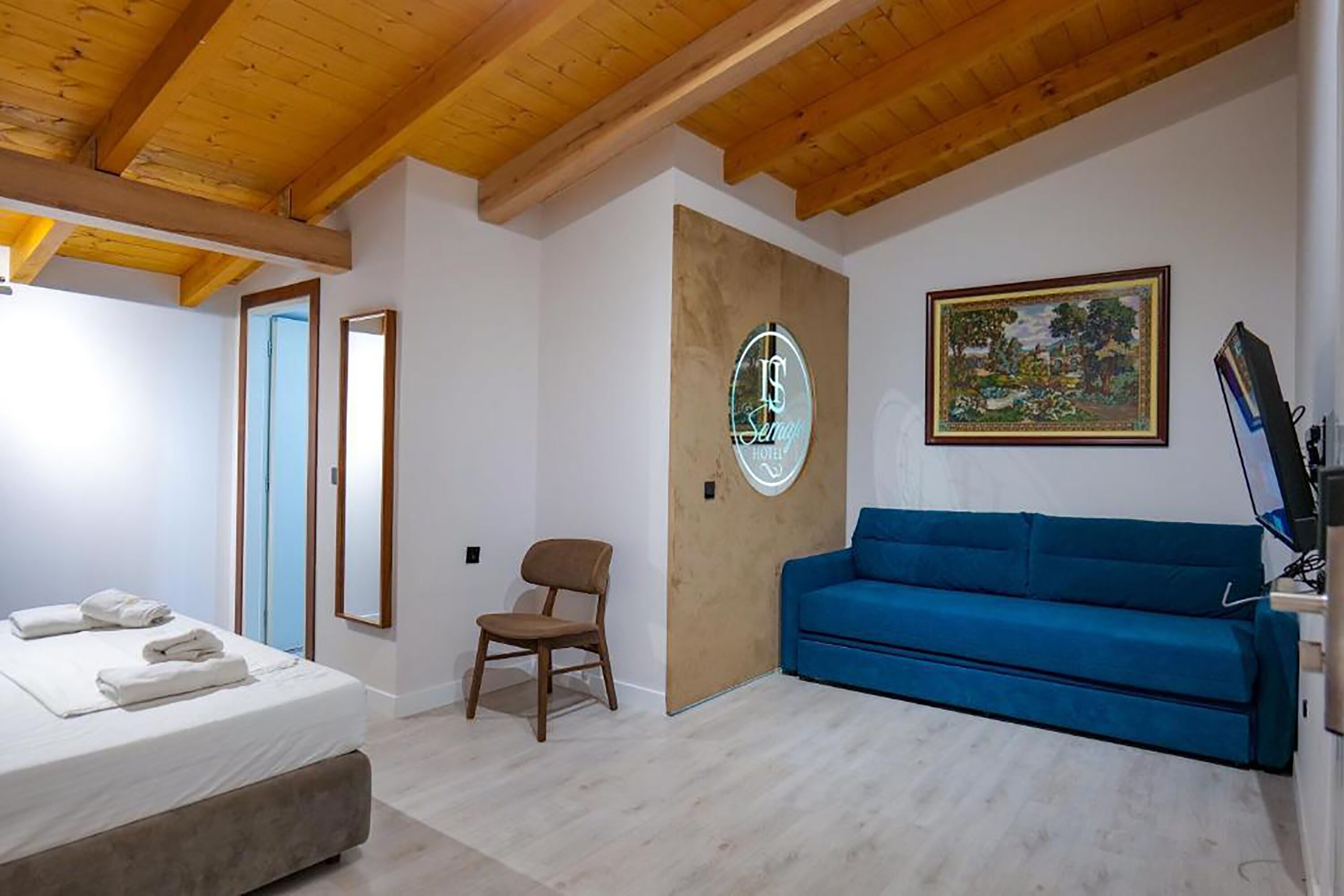 A hotel room with a bed, a blue sofa, a wooden chair, a painting, and a sign on the wall.