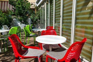 Une terrasse extérieure avec des tables et des chaises rondes, dont certaines sont de couleur rouge et verte, et un mur de verre en arrière-plan.