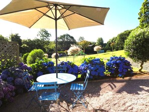 An outdoor patio with a round table and blue chairs, surrounded by a garden with hydrangeas and a pergola. - "Le Macareux" Vacation Rental & Chalet: Lush, Green Setting—Beach Within Walking Distance (Trévou-Tréguignec)