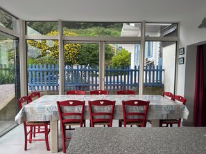 Un coin repas avec une table dressée pour six personnes, entouré de chaises rouges, de grandes fenêtres et d’une clôture bleue visible à travers le verre.