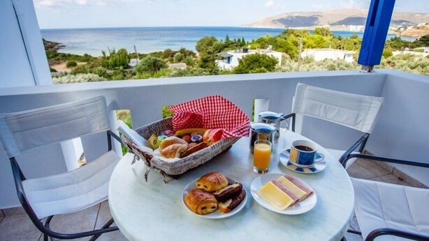 A balcony with a table set for breakfast, including croissants, a basket of fruits, and a glass of orange juice. - Villa Maniati Studios (Andros)