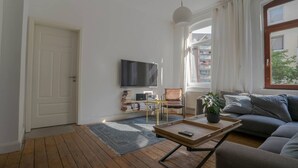 Un salon avec un plancher en bois, un canapé, une table basse, un téléviseur, une bibliothèque et une fenêtre avec des rideaux.