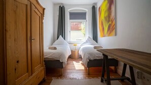 Une petite chambre agréable, dotée de deux lits simples, d’une armoire en bois, d’une table et d’une fenêtre avec des rideaux.
