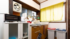A compact kitchen with a microwave, kettle, and a window with yellow curtains. There are two trash bins and a shelf with kitchen utensils. - Tokyo House (Tokyo)