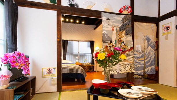 A traditional Japanese room with tatami flooring, a futon bed, a wooden TV stand with a pink orchid, and a table set with plates and bowls. - Tokyo House (Tokyo)
