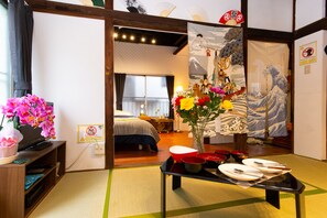 A traditional Japanese room with tatami flooring, a futon bed, a wooden TV stand with a pink orchid, and a table set with plates and bowls. - Tokyo House (Tokyo)