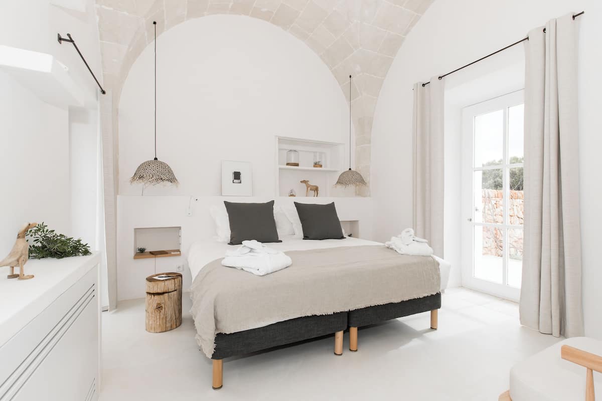 A modern bedroom with a large bed, two bedside tables, a small wooden stool, and a large window with curtains.