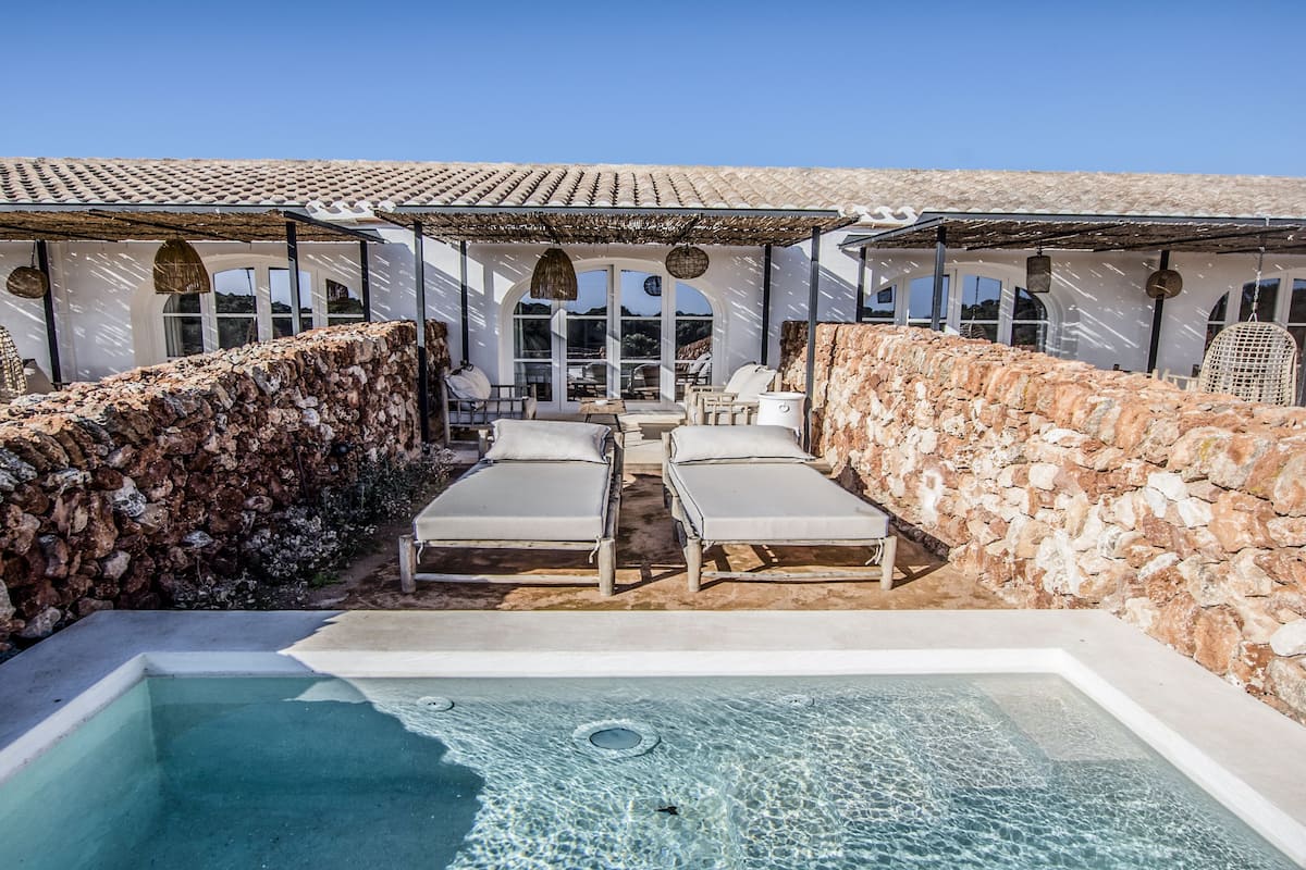 A pool area with lounge chairs and a hot tub, surrounded by stone walls and covered seating areas.