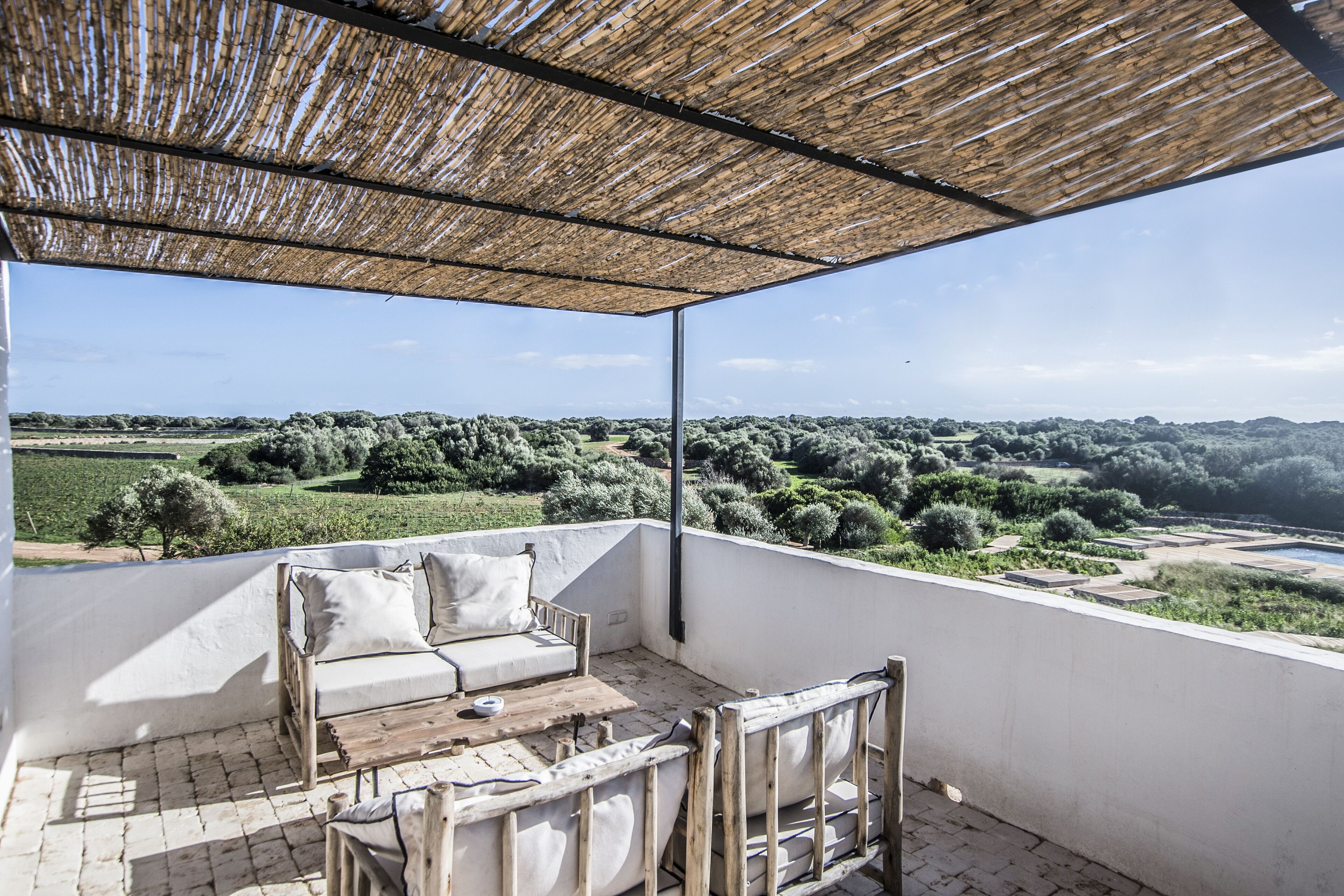 Un balcon abrité, doté d’un canapé blanc et de chaises en bois, qui offre une vue sur un paysage luxuriant, avec des arbres et des champs.