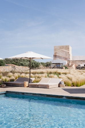 Zona de piscina con sillones, una sombrilla blanca y un edificio de piedra al fondo.