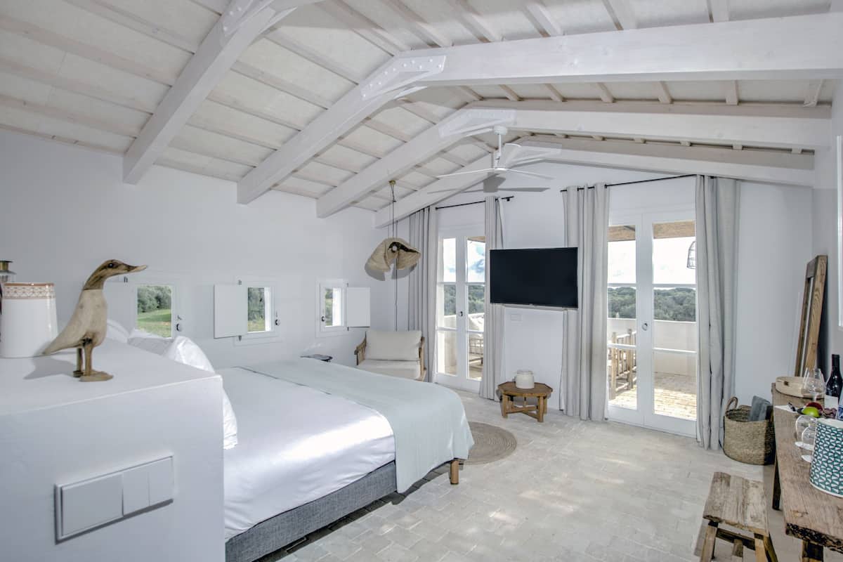 A spacious bedroom with a large bed, a ceiling fan, a TV, and a balcony with a view of the outdoors.