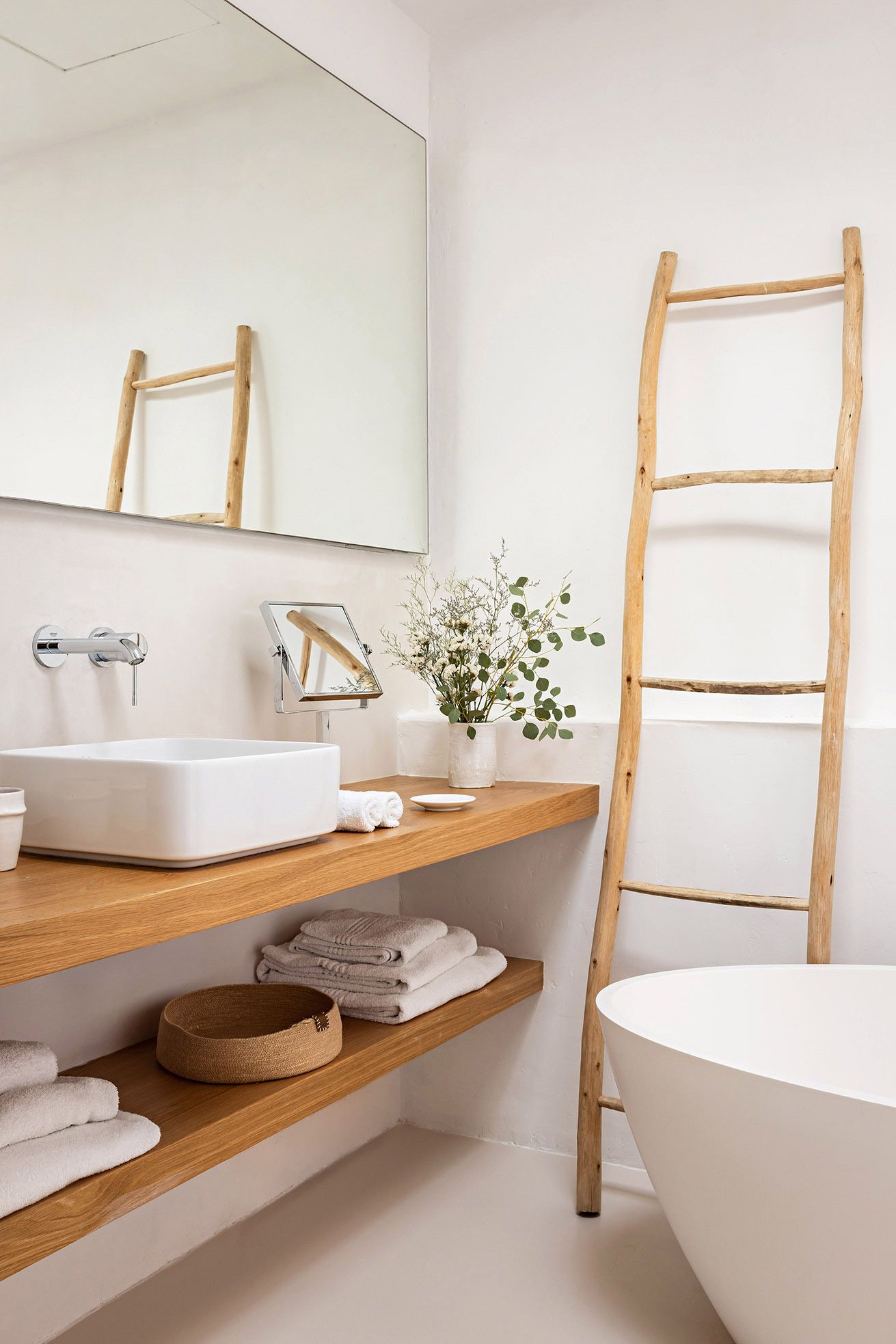 A bathroom with a wooden vanity, a white sink, a mirror, a wooden ladder, a white bathtub, and a shelf with towels and a basket.