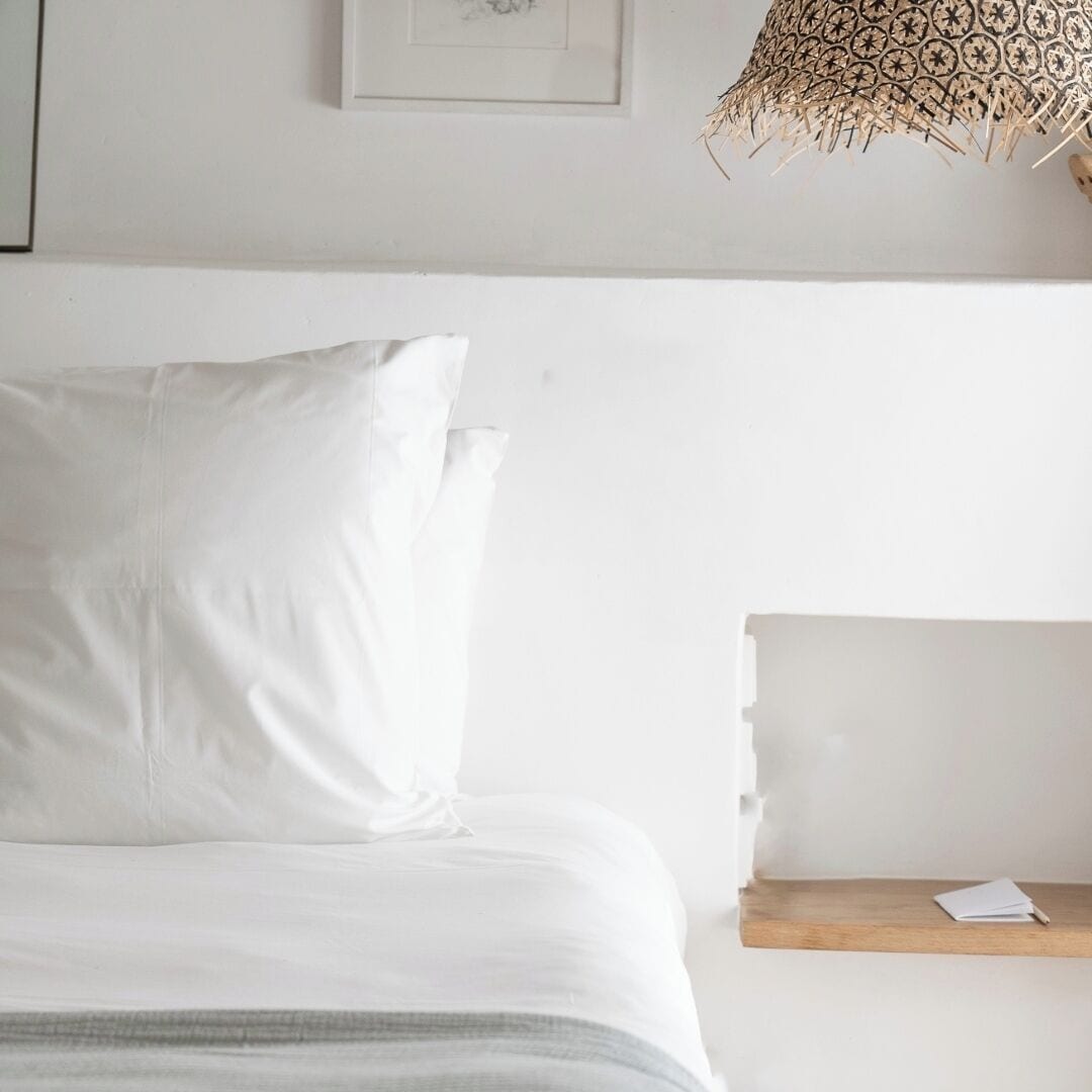 Una cama bien tendida con ropa de cama blanca, una mesita de noche de madera con un libro y una lámpara colgante con un diseño decorativo.