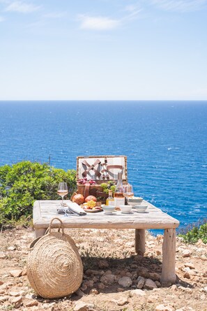 A picnic setup with a wicker basket, a wooden table, and a view of the sea. - Torre Vella – Fontenille Collection (Alayor)