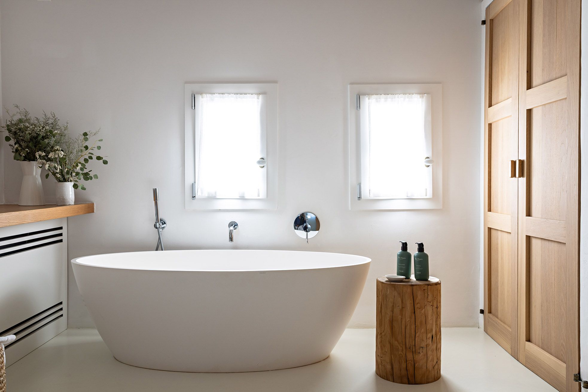 A modern bathroom with a freestanding bathtub, two windows, a wooden stool, and a wooden door.