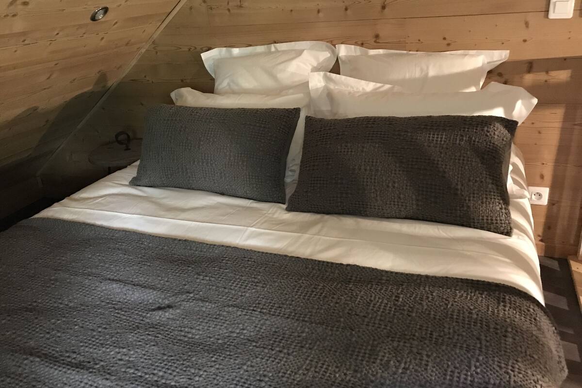 Une chambre à coucher confortable, aménagée en mezzanine, avec un plafond en pente, des murs en bois et un lit recouvert d’une literie blanche et gris foncé.
