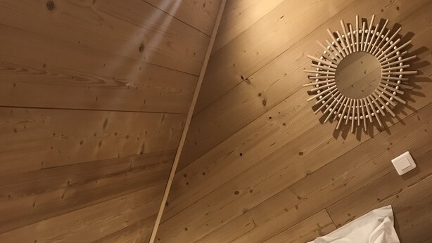 Un plafond en bois avec un luminaire circulaire et un lit recouvert d’une literie blanche et agrémenté de deux coussins de couleur moutarde.