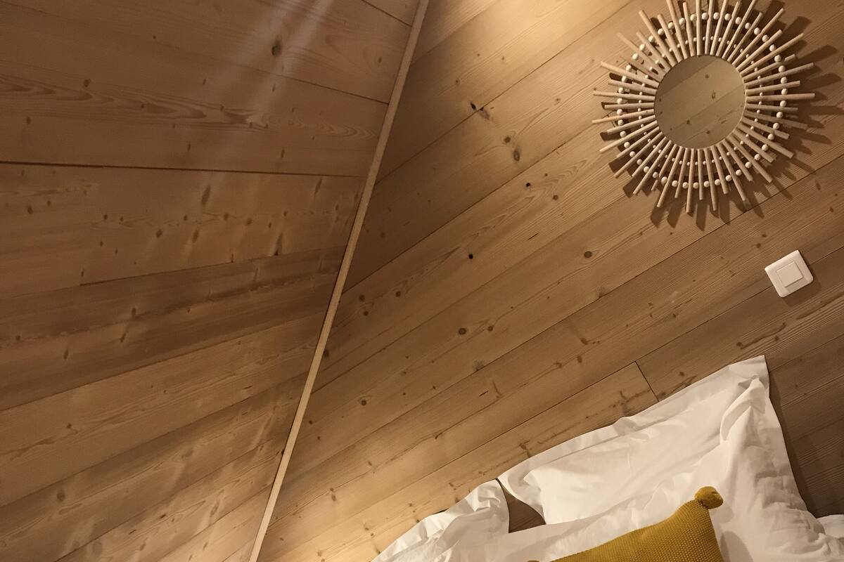 Un plafond en bois avec un luminaire circulaire et un lit recouvert d’une literie blanche et agrémenté de deux coussins de couleur moutarde.