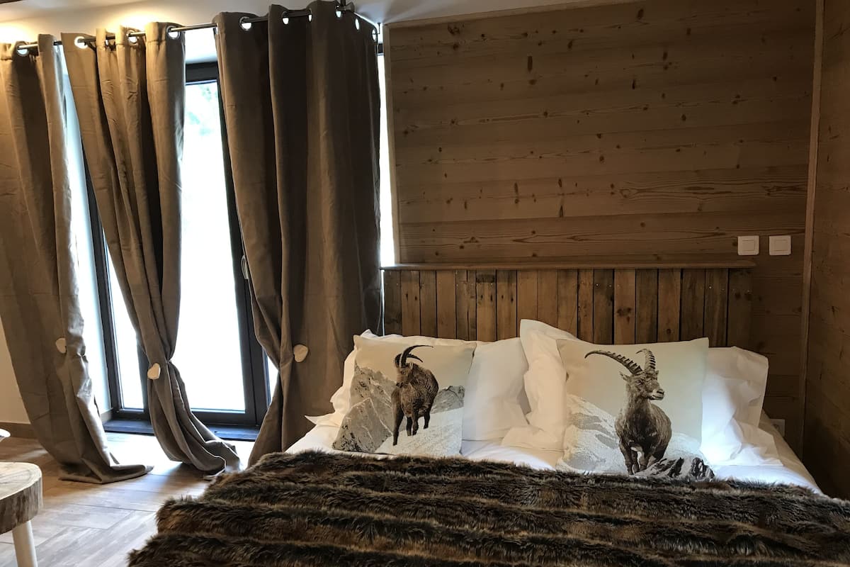 Une chambre à coucher avec une tête de lit en bois, un grand lit avec une literie blanche et deux coussins à motifs de chèvres, un plaid en fausse fourrure et une fenêtre avec des rideaux.