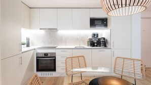 Ett modernt kök med vita skåp, en inbyggd ugn, en mikrovågsugn och en kaffemaskin. Det finns två fåtöljer i rotting och en hängande lampa.
