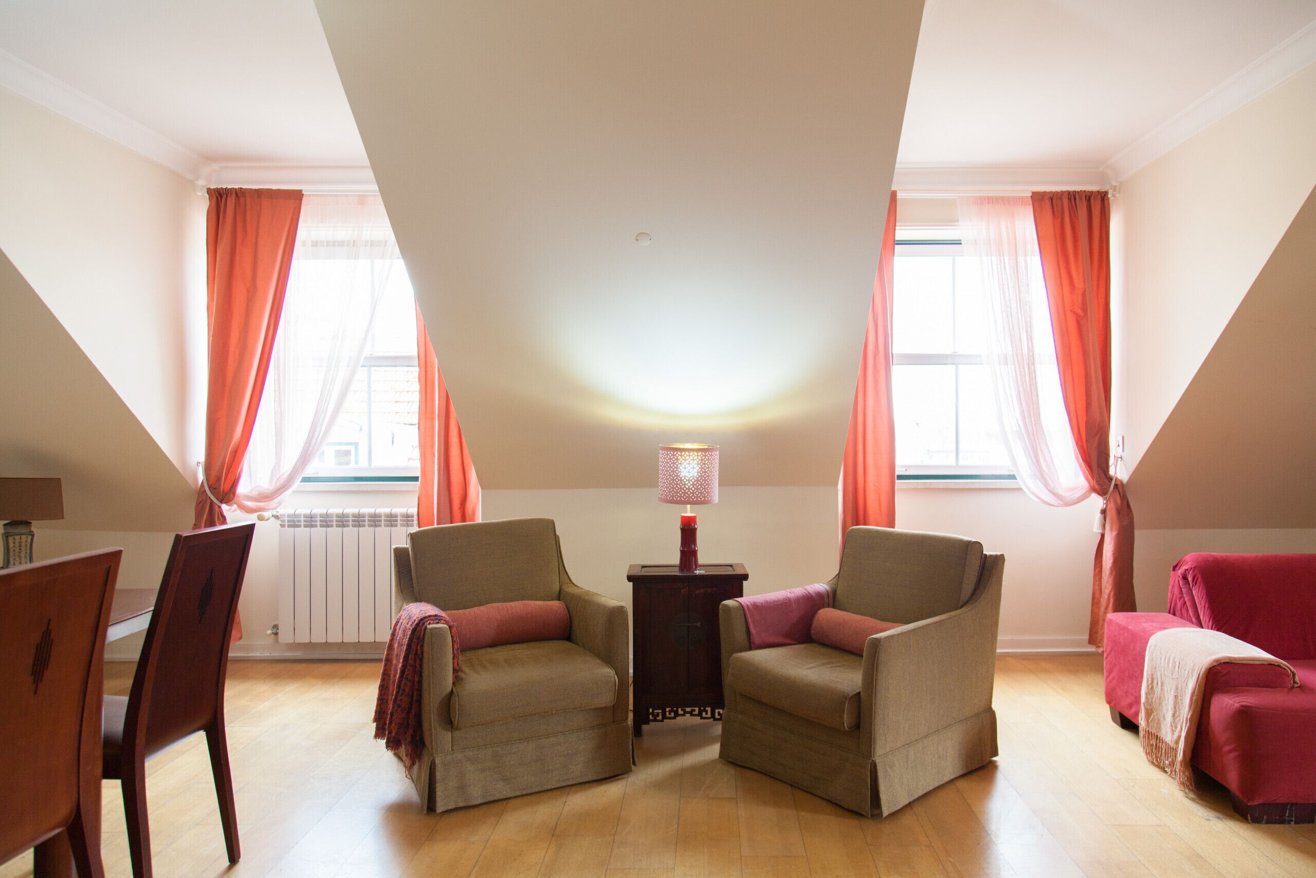 Ein Zimmer mit zwei Sesseln, einem kleinen Tisch mit Lampe, einem roten Sofa und einem Esstisch mit Stühlen.
