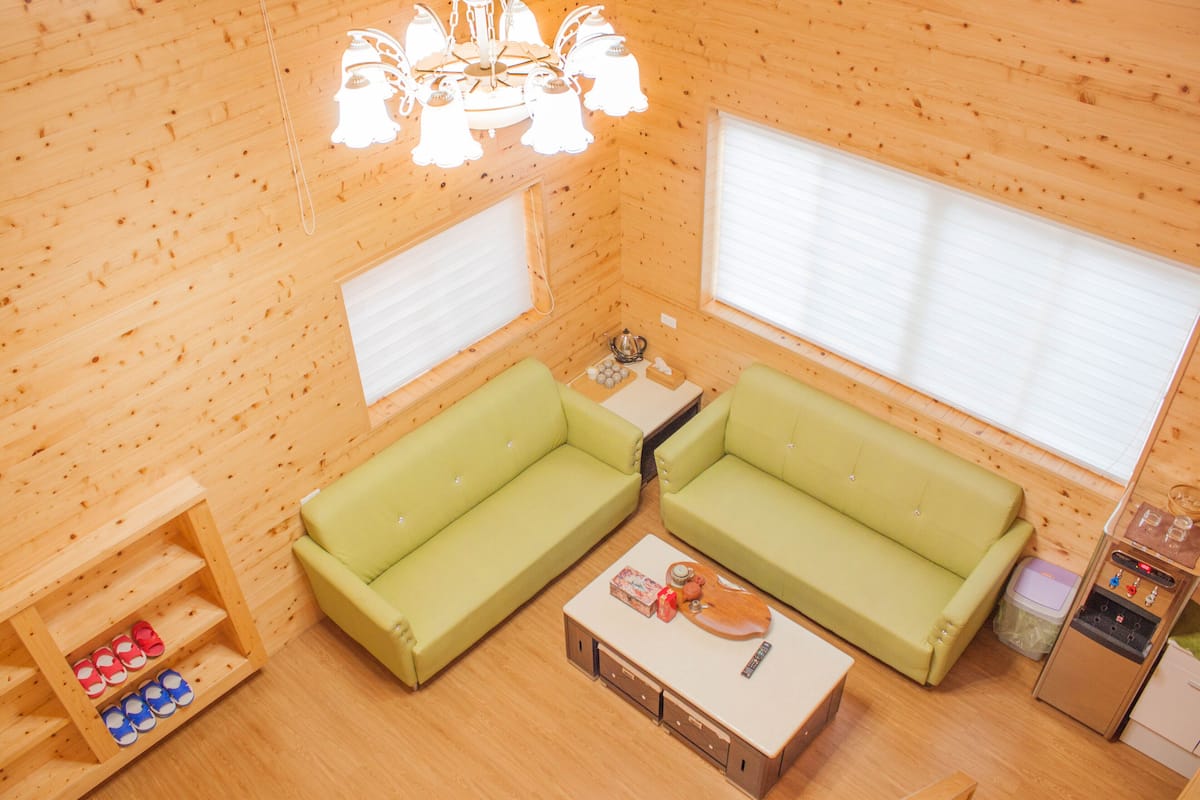 Una habitación acogedora con dos sofás verdes, una estantería de madera con zapatos, una mesita con una tetera y una ventana con persianas.