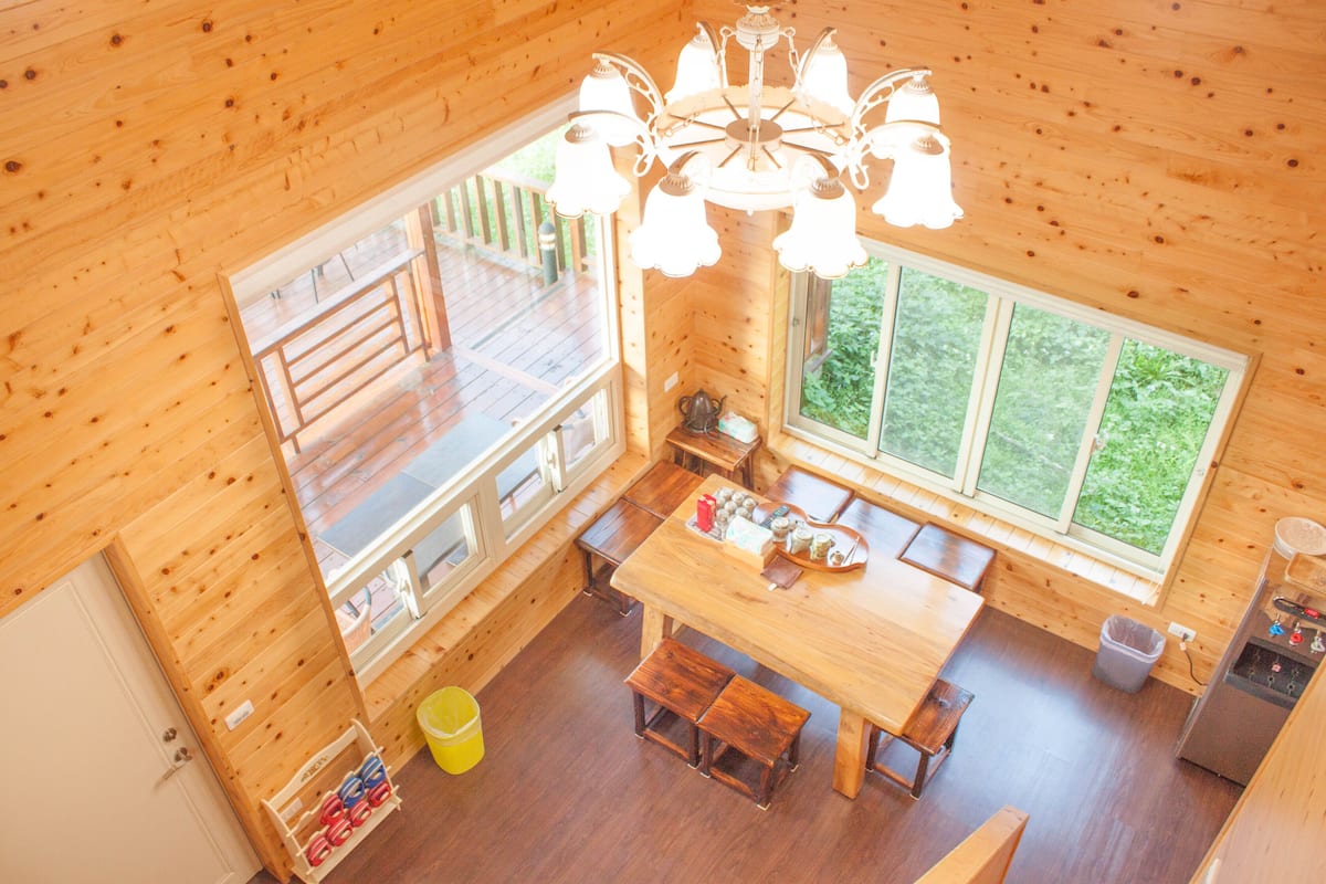 Interior de una cabaña de madera con comedor, ventanas grandes y un candelabro.