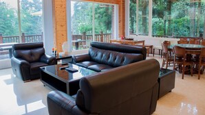 A spacious living area with a leather sofa, a coffee table, and a dining table with chairs. Large windows provide a view of the surrounding greenery.