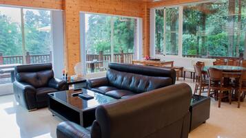 A spacious living area with a leather sofa, a coffee table, and a dining table with chairs. Large windows provide a view of the surrounding greenery.