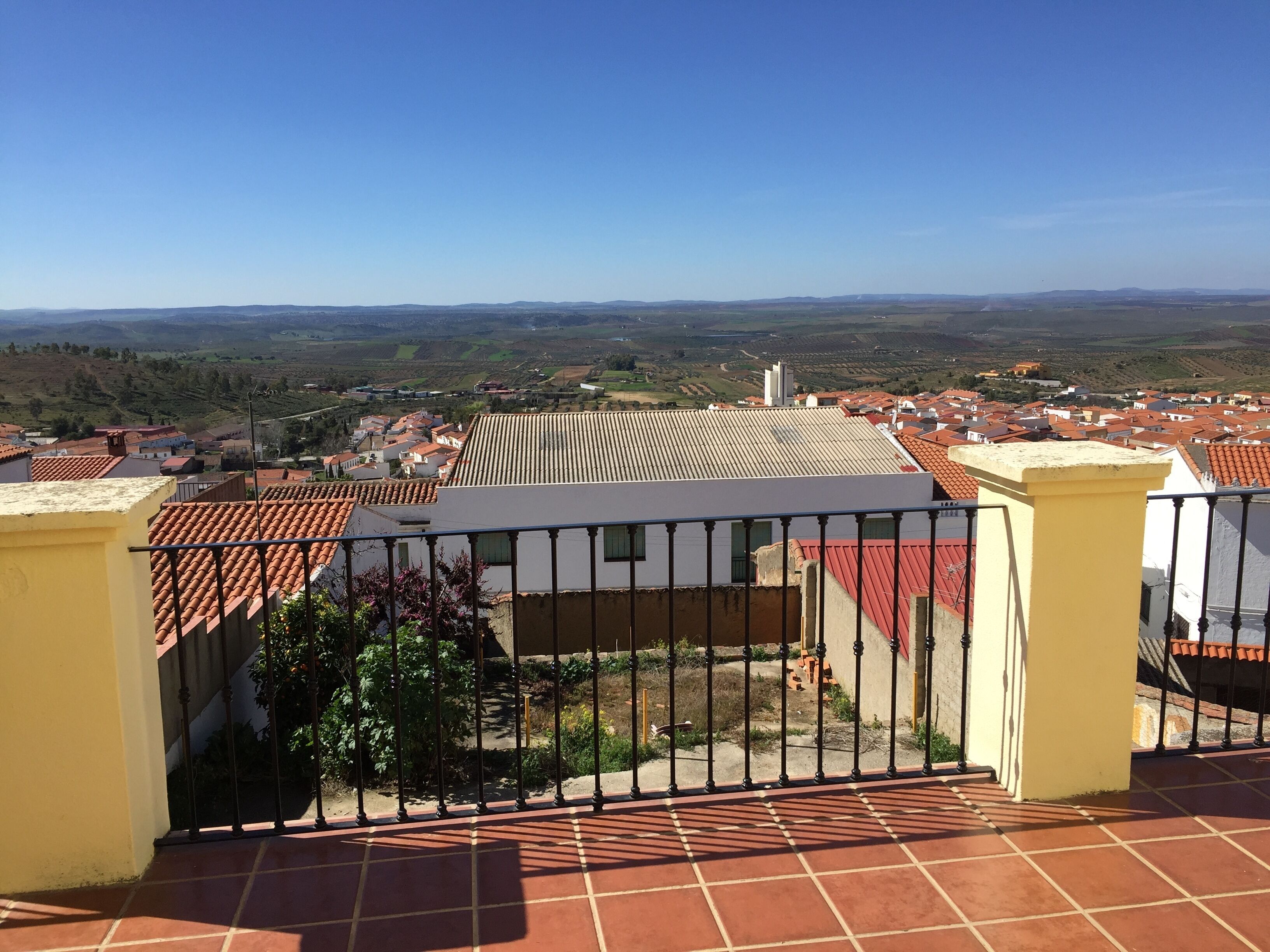 A terrace with a tiled floor and a metal railing, overlooking a town with red-roofed buildings and green hills in the distance.