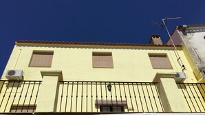 Un edificio amarillo con tres ventanas, un balcón con barandillas negras y una antena en el techo.