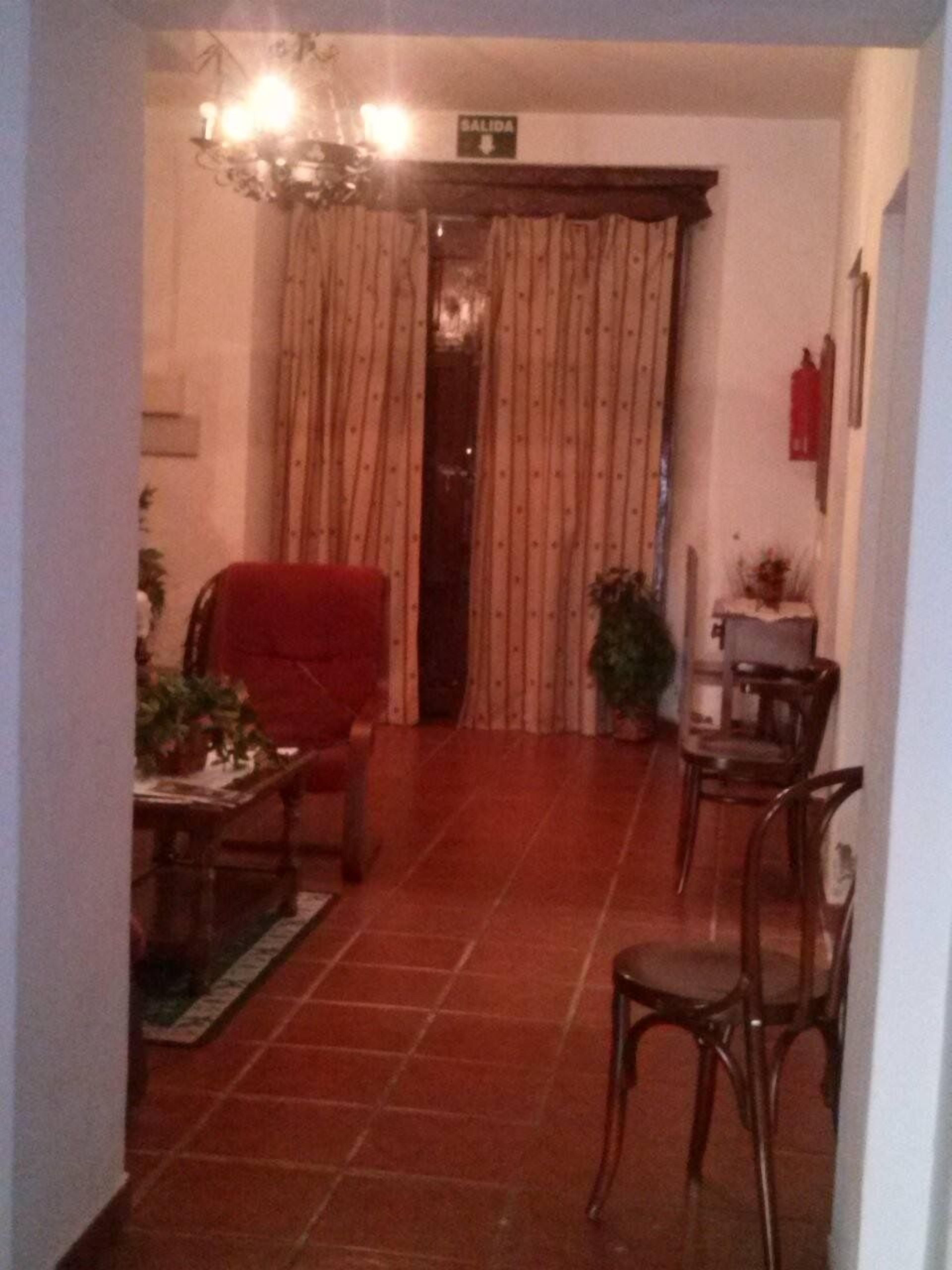Una habitación con un sillón rojo, una mesa de centro de madera y una mesa de comedor con sillas. Hay una planta en maceta y una mesita con un jarrón. El suelo está cubierto de baldosas y hay una puerta con cortinas.