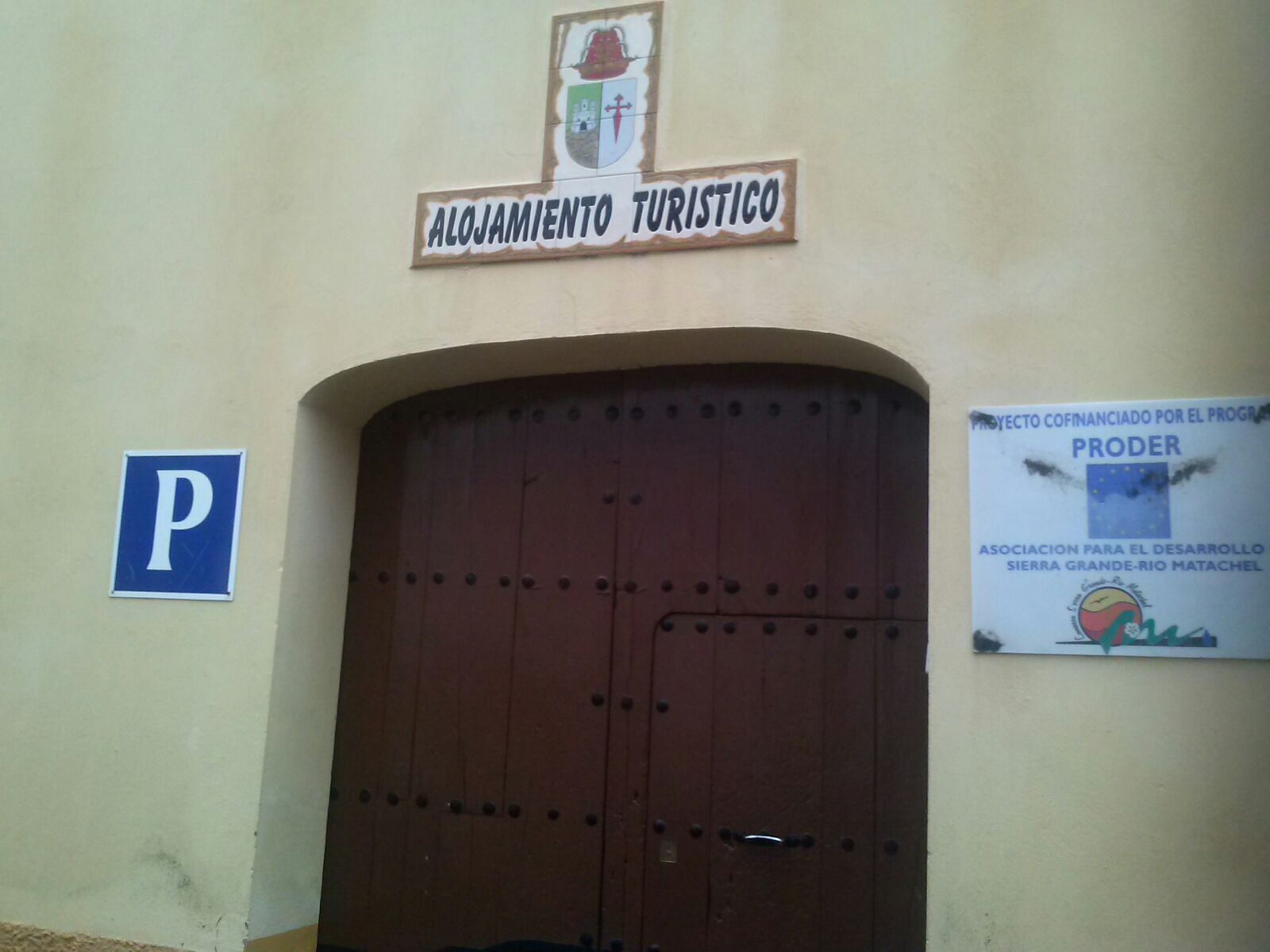 Entrada de un edificio con un amplio puerta de madera, un letrero que dice 'Alojamiento Turistico' en la parte superior y un cartel de estacionamiento a la izquierda.