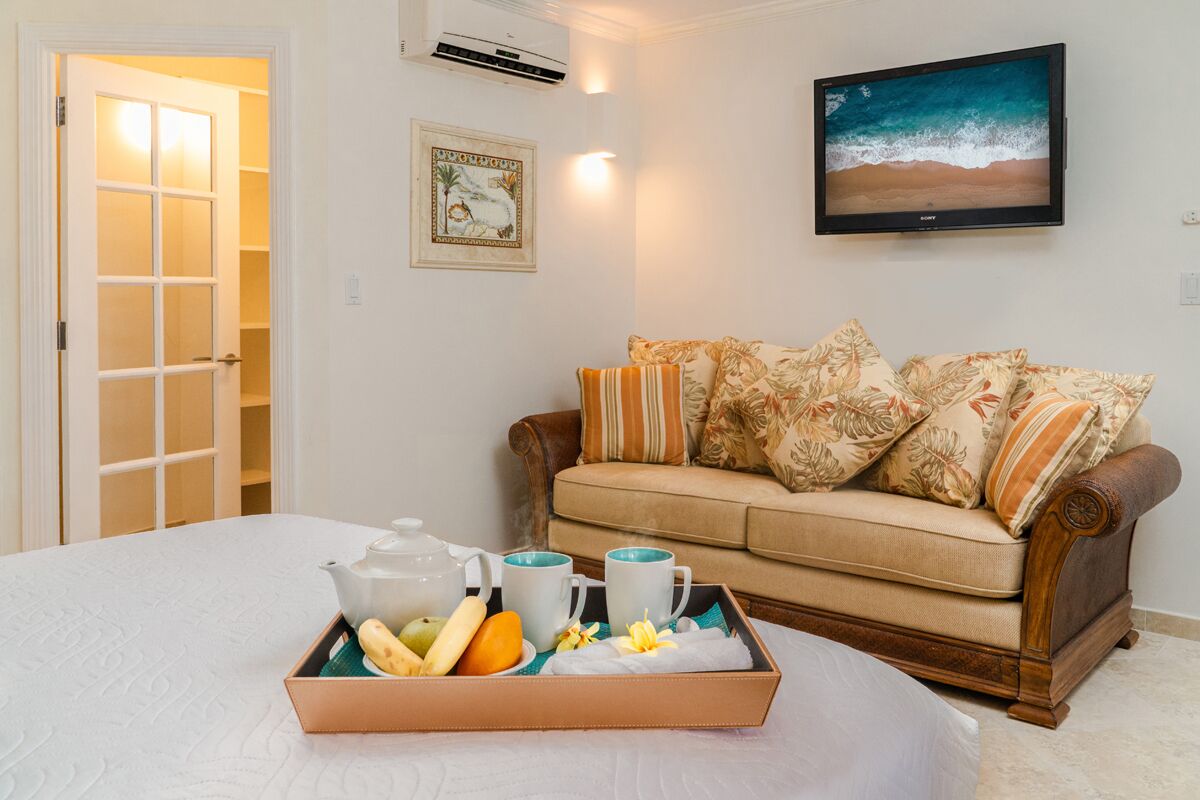 Una habitación con una cama, un sofá de madera, una bandeja con alimentos para el desayuno, un televisor montado en la pared y un cuadro enmarcado.