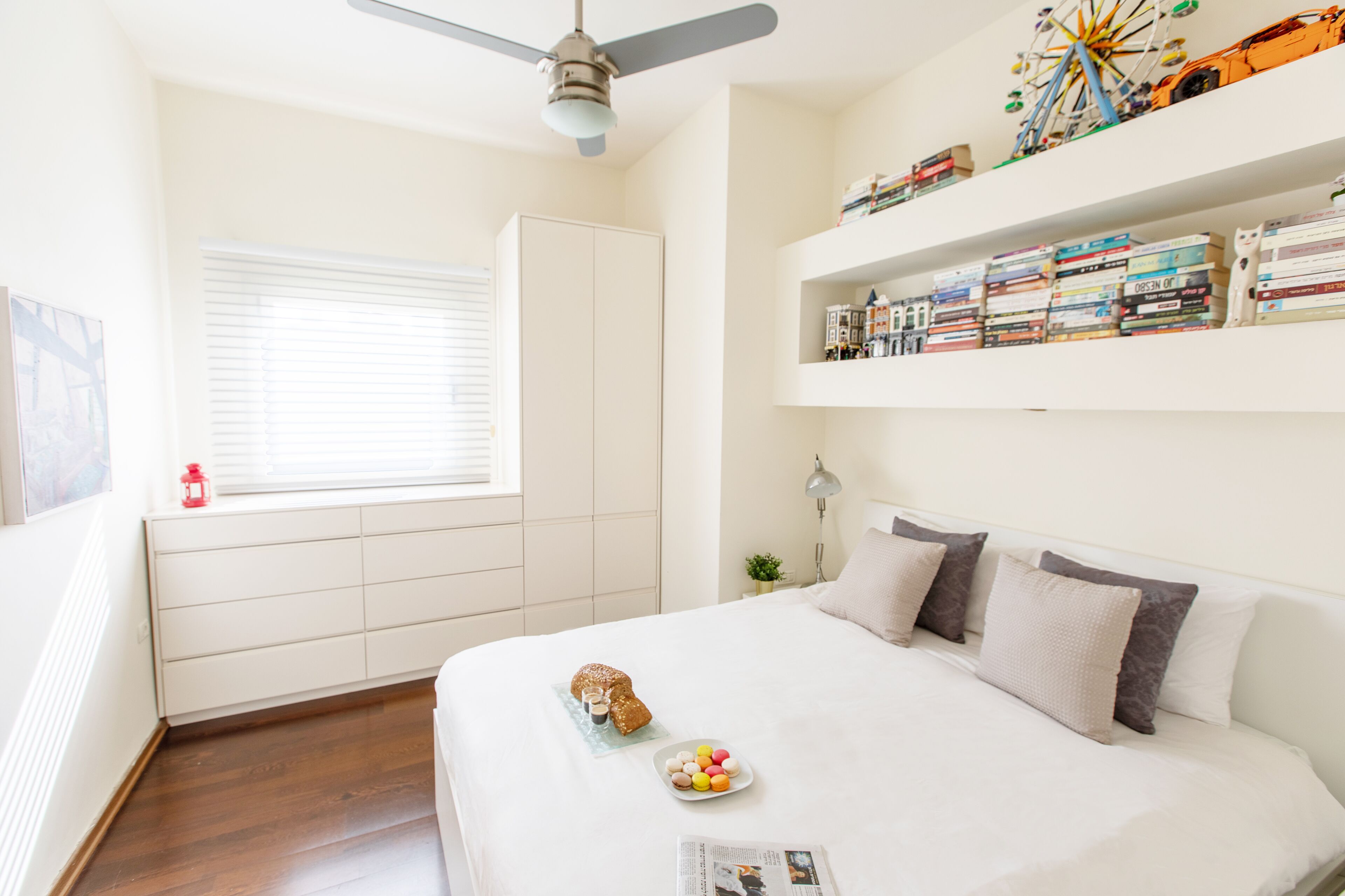 A bedroom with a bed, a ceiling fan, a window with blinds, a bookshelf, and a small plant.