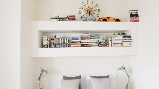Ein ordentlich eingerichtetes Schlafzimmer mit einem Bett, zwei Nachttischlampen, einer kleinen Pflanze und einem Bücherregal mit Büchern und einem Spielzeugauto.