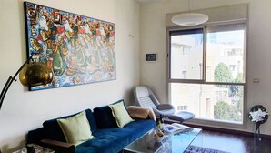 Ein Wohnzimmer mit einem blauen Sofa, einem Glastisch, einem großen abstrakten Gemälde, einem modernen Sessel und einem großen Fenster mit Blick auf Gebäude.