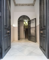 A hallway with a white tiled floor, a black ornate door with a brass doorknob, and a decorative ceiling medallion.