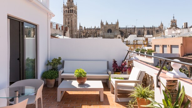 Una terraza en la azotea con un banco blanco, una mesa de vidrio y plantas en macetas. Se observa un edificio histórico con una torre al fondo.