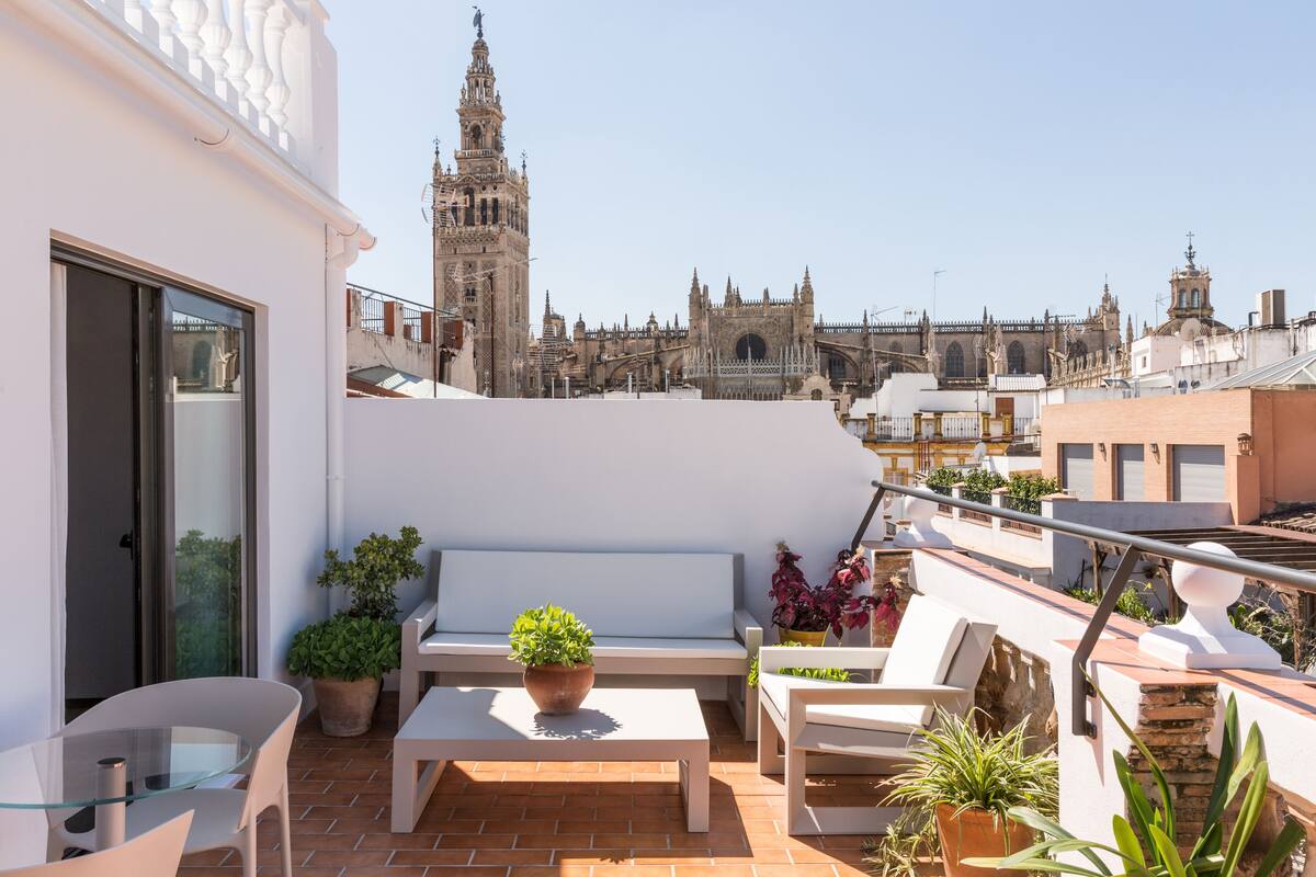 A rooftop terrace with a white bench, a glass table, and potted plants. A historic building with a tower is visible in the background.