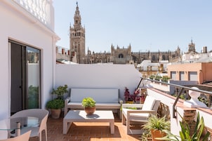A rooftop terrace with a white bench, a glass table, and potted plants. A historic building with a tower is visible in the background.