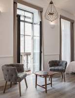 A spacious room with large windows, two tufted armchairs, a small wooden table, and a hanging light fixture.