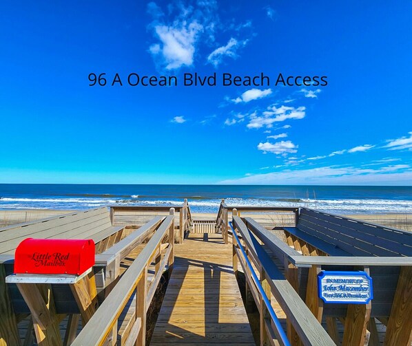 A wooden boardwalk leading to a beach with a mailbox labeled "Little Red Mailbox" and a sign commemorating John Macomber.