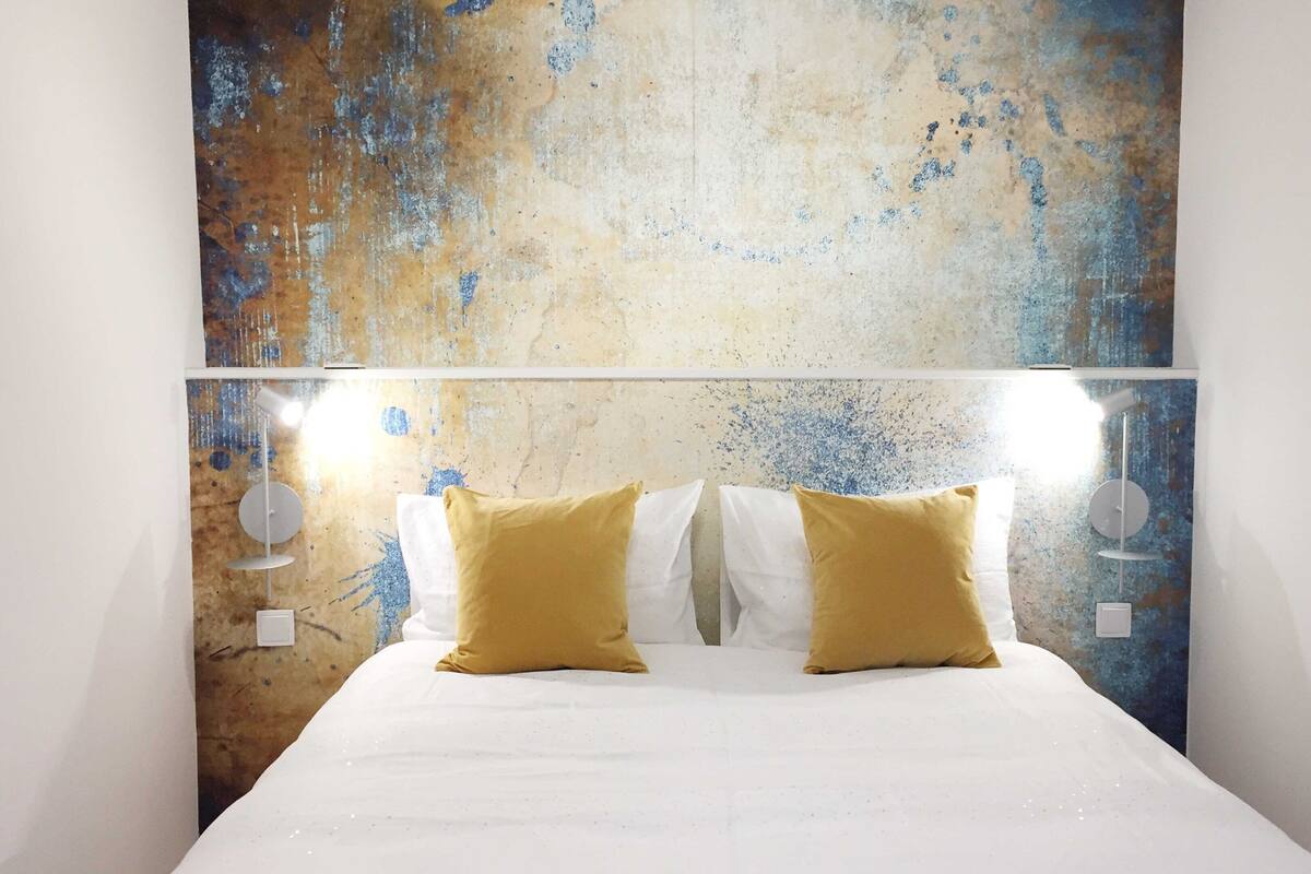 Un lit avec une literie blanche et deux coussins de couleur moutarde, placé devant un mur texturé aux tons bleus et beiges.
