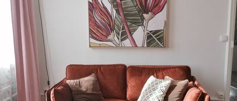 A living room with a red velvet sofa, a round coffee table, and a large floral painting on the wall.