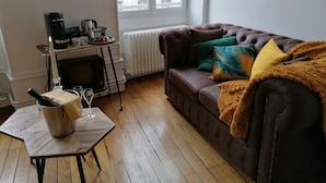 A cozy living room with a brown leather sofa, a small wooden table with a candle, and a window with a view of buildings. - B&B 43 4 (Le Puy-en-Velay)
